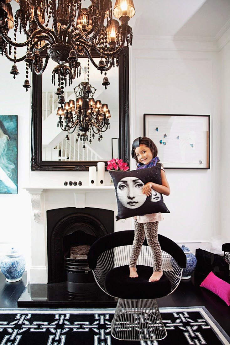 Kleines Mädchen mit Kissen auf Klassikerstuhl, offener Kamin mit Spiegel, Kronleuchter - traditionelles Ambiente mit schwarzen und weissen Farbakzenten