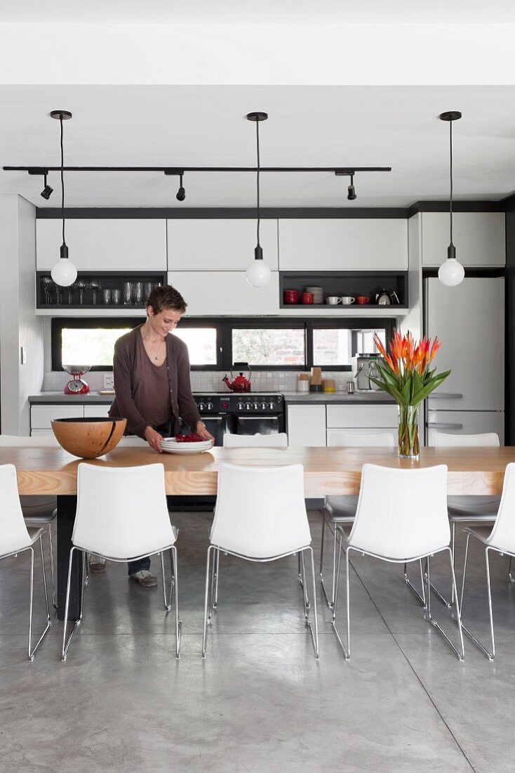 Frau an langem Esstisch mit Schalenstühlen und einfachen Hängeleuchten vor einer modernen Einbauküche