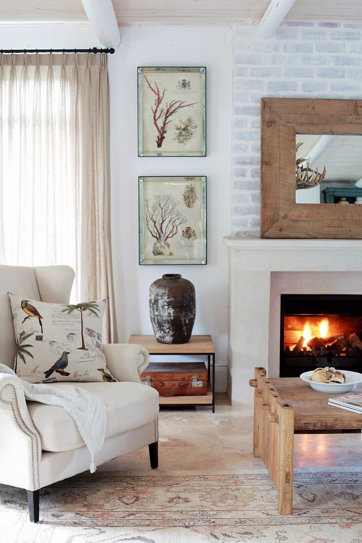 Wohnzimmer in Country-Style mit Ohrensessel und Couchtisch vor Kaminfeuer und weiss getünchter Ziegelwand