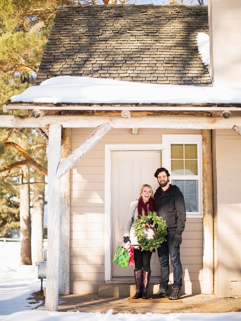 Junges Paar steht mit Tannenkranz vor winterlichem Haus