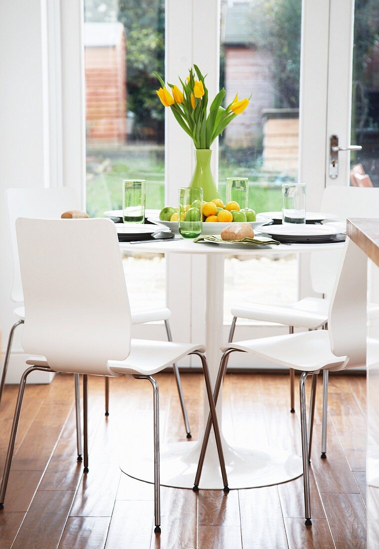 weiße Schichtholzstühle an gedecktem, rundem Tisch mit gelbem Tulpenstrauss; Fenstertür zum Garten im Hintergrund