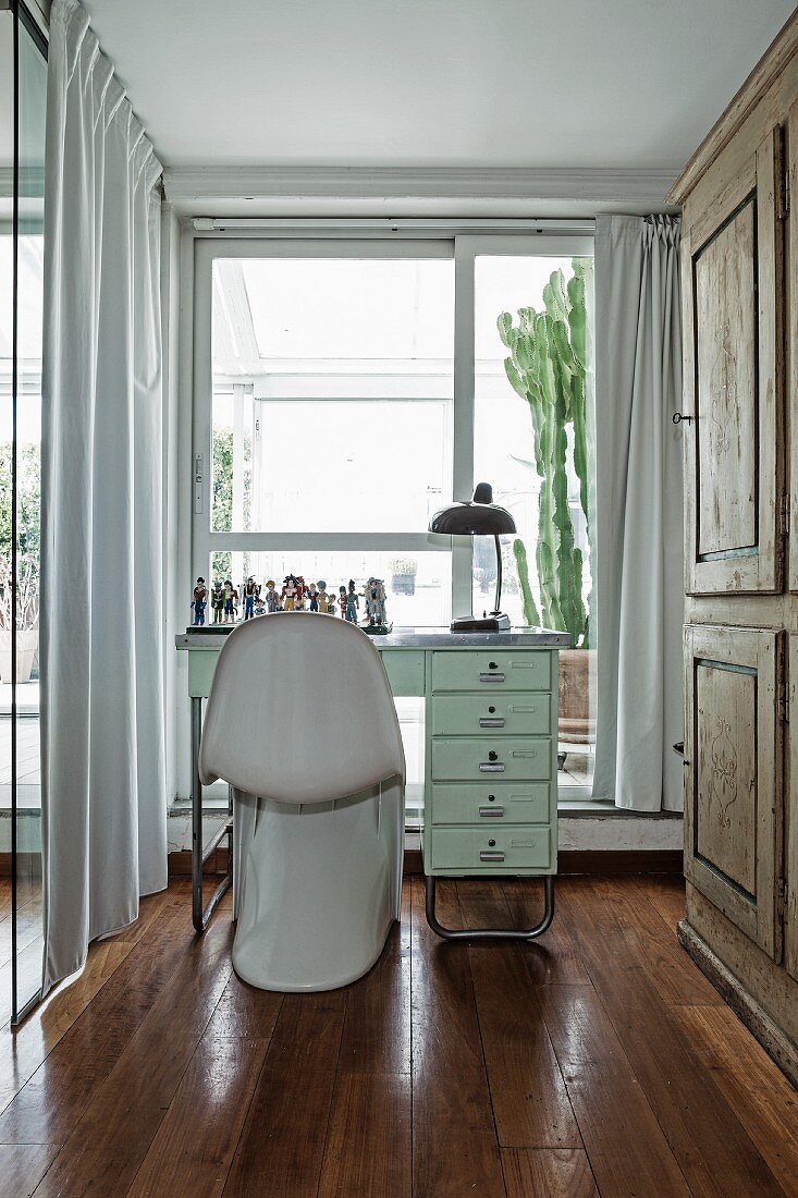 Weisser Klassiker Schalenstuhl vor Schreibtisch in schmalem Raum mit bodenlangen Vorhängen vor Fenster
