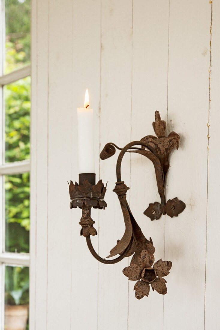 Brennende Kerze in Metall Wandkerzenleuchter mit Blumenelementen im Vintage Stil, an heller Holzwand