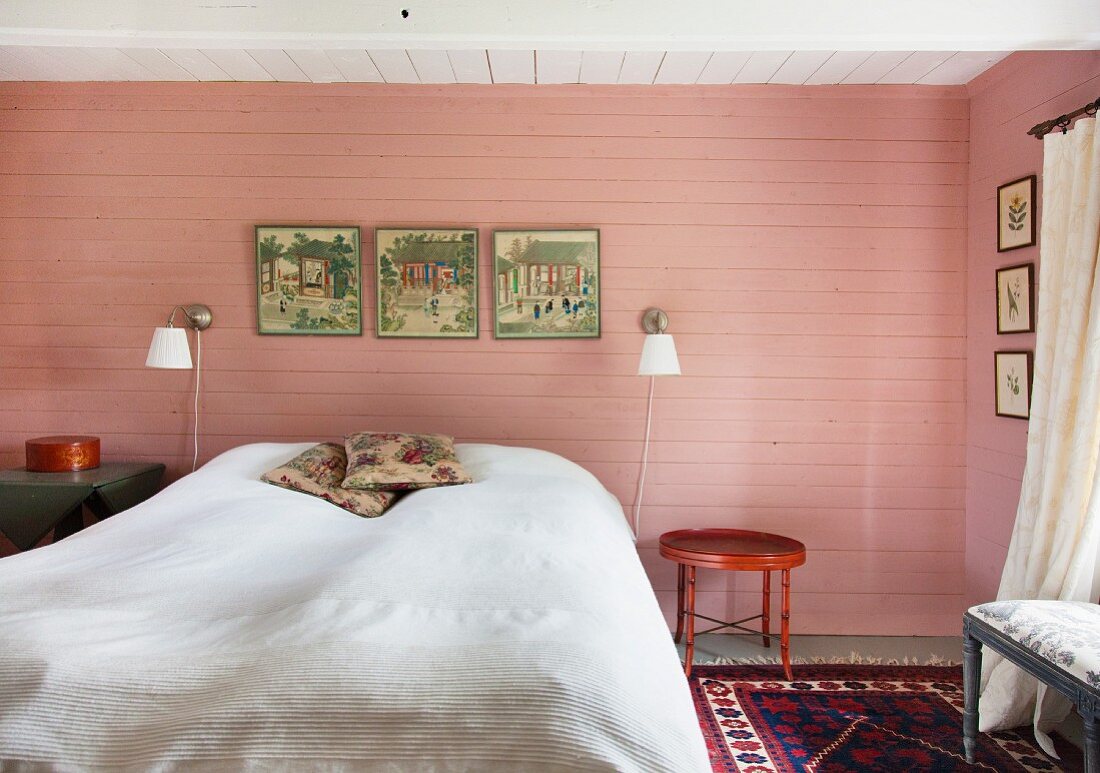 Doppelbett mit weisser Tagesdecke vor rosa getönter Holzwand, mit Wandleuchten, in ländlichem Schlafzimmer