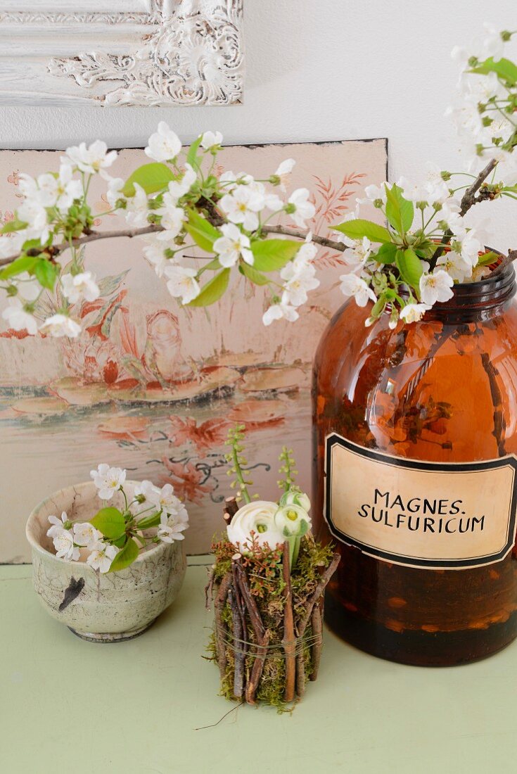 Keramikschale mit Wildkirschblüten, Ranunkel in mit Ästen umwickeltes Gläschen und Apothekerflasche mit Wildkirschblüten vor antikem Emailleschild