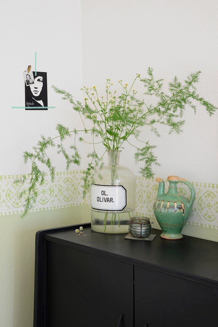 Alte Apothekerflasche mit Zierspargel und Wiesenblumen neben grünem Vintage Keramikkrug und antikes Teelicht auf schwarzem Sideboard, an Wand grüne Bordüre