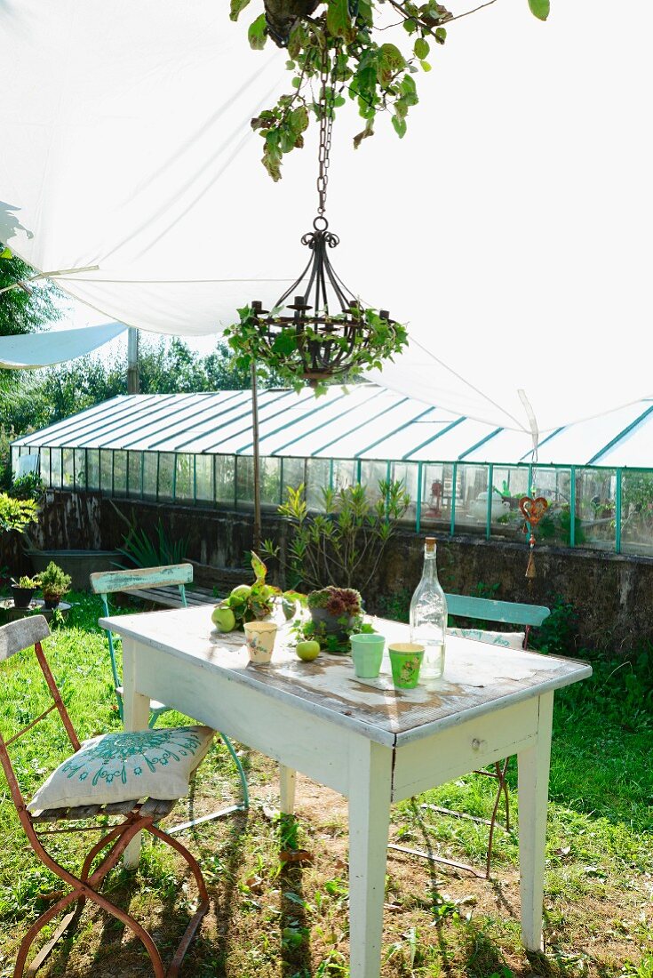 Alte Gartenstühle mit Kissen um rustikalen Gartentisch mit Bechern und Vintage Limonadenflasche unter Efeu umranktem Kronleuchter und Sonnensegel, im Hintergrund Gewächshaus