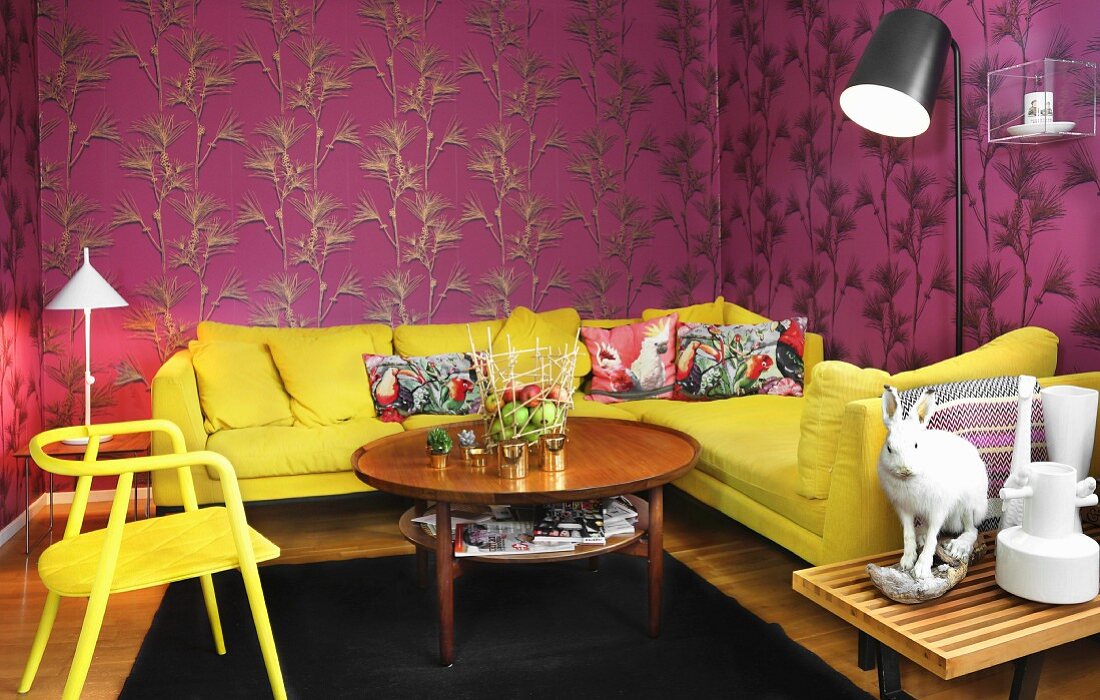 Yellow corner sofa with colourful scatter cushions, table and chair against mauve wallpaper