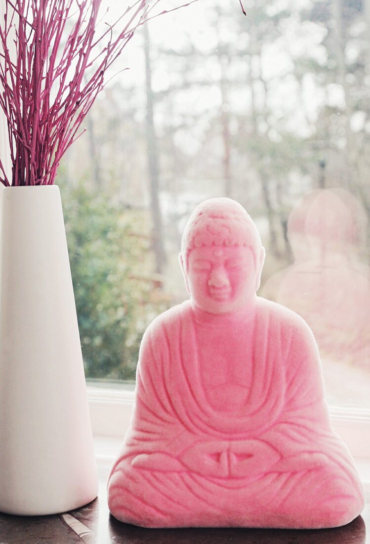 Rosafarbene Buddhafigur neben weißer Vase mit roten Zweigen auf Fenstersims