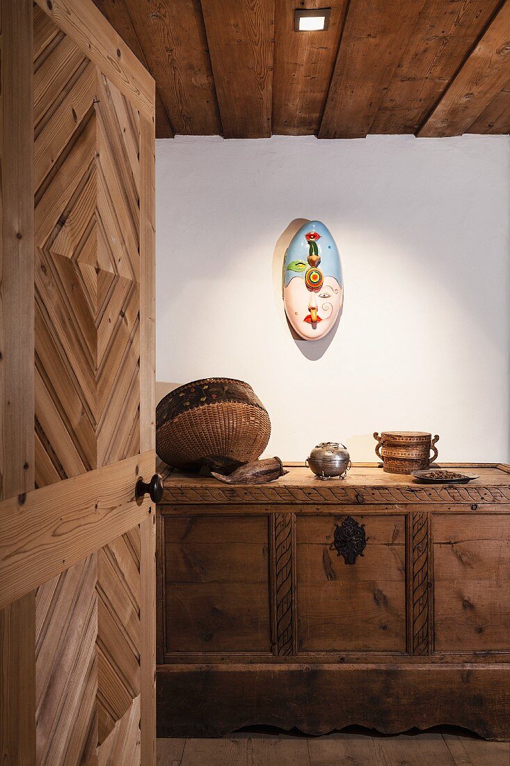 Blick durch geöffnete Holztür auf bemalte moderne Wandskulptur und geschnitzte Bauerntruhe mit Deko