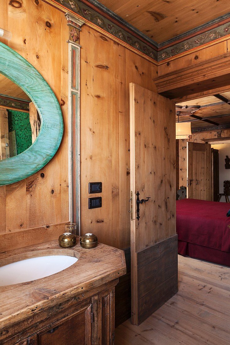 Rustikaler Waschtisch und Ovalspiegel mit türkis lasiertem Holzrahmen; Blick durch die offene Badtür in den Schlafraum