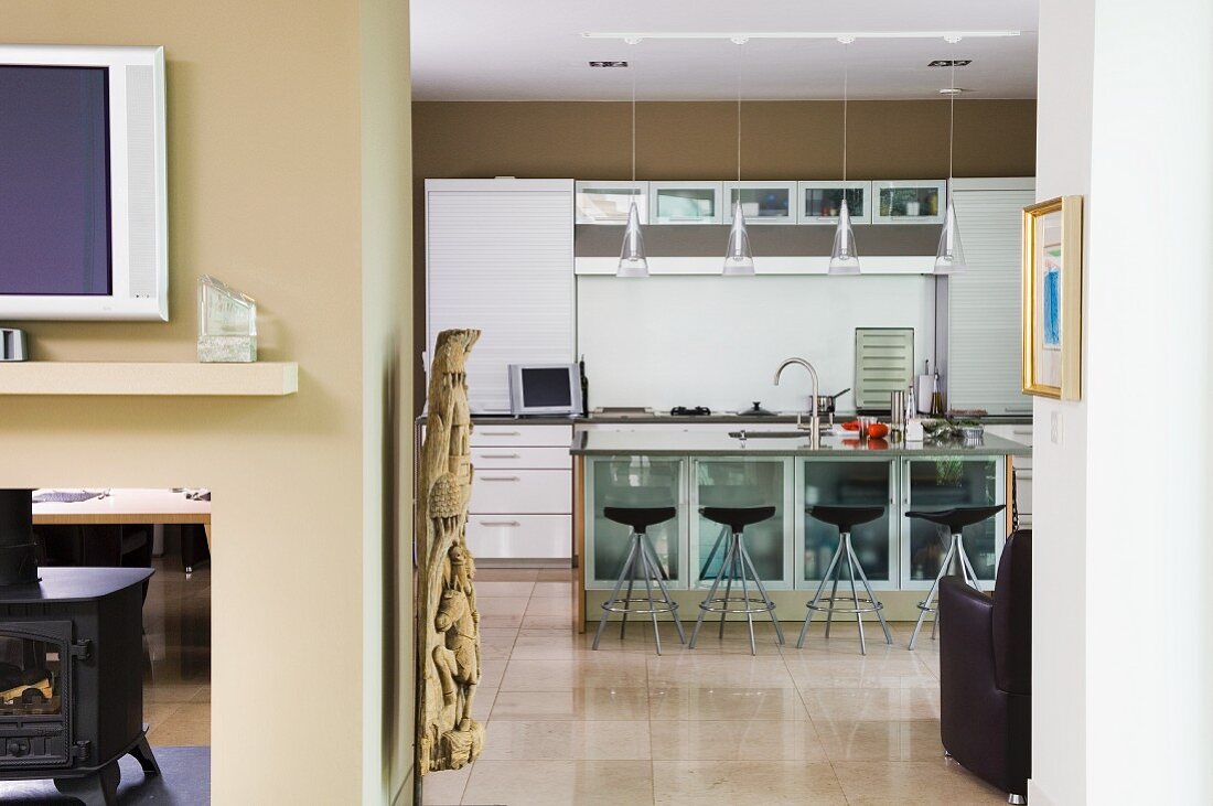 Blick in offene Küche mit sandfarbenen Wänden, Barhocker mit schwarzen Sitzschalen vor Theke mit Glastüren an Unterschränken