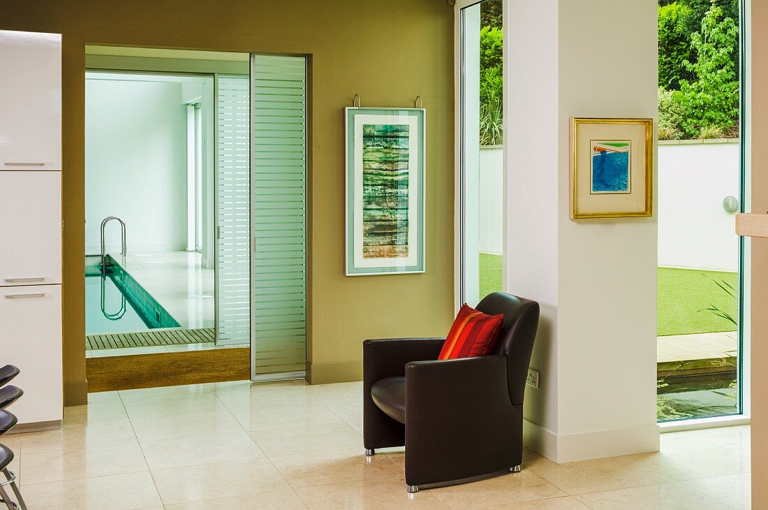 Brauner Ledersessel vor Eckpfeiler, zwischen raumhohen Fenstern mit Gartensicht, seiltich offene Schiebetüren und Blick auf Indoor Pool