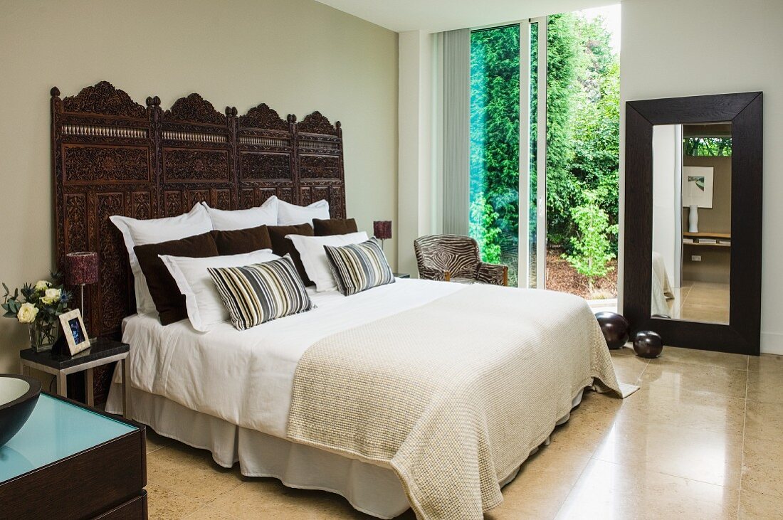 Doppelbett mit Tagesdecke, Kissenstapel vor geschnitzen, indischen Holzpaneelen an sandfarbener Wand, im Hintergrund Glas Schiebetüren mit Gartenblick
