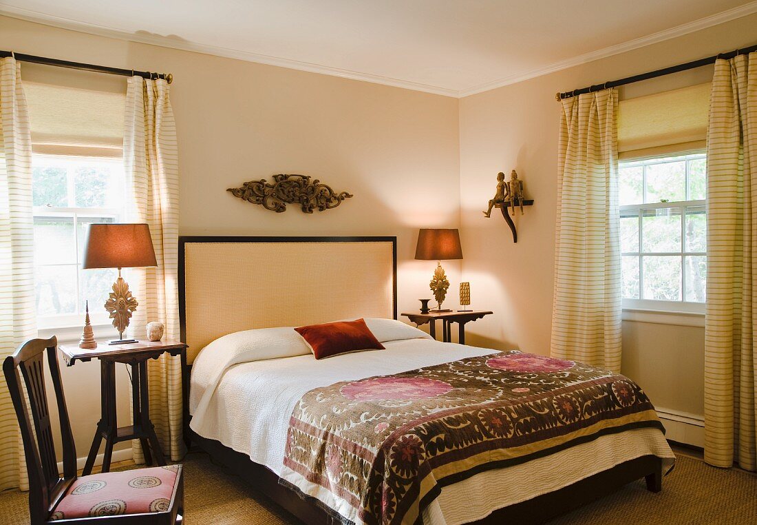 Gemusterte Tagesdecke auf Doppelbett mit hohem Kopfteil, in Zimmerecke zwischen Sprossenfenster mit bodenlangen Vorhängen