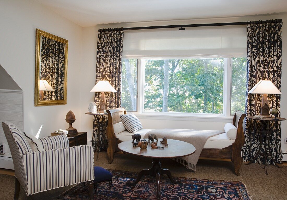 Antikes Schlittenbett vor Fenster mit gemusterten, bodenlangen Vorhängen, gegenüber Sessel mit gestreiftem Bezug vor rundem Tisch aus dunklem Holz