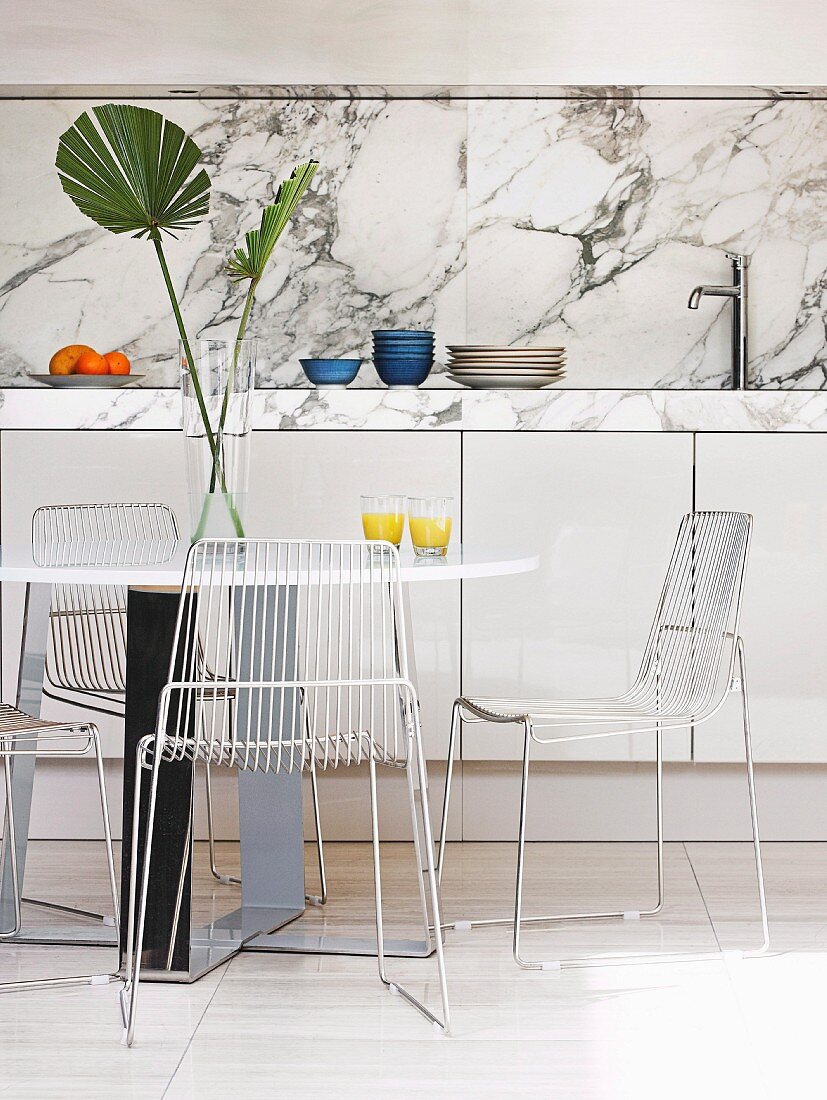 Esstisch, Drahtstühle und Palmenblatt-Deko vor weisser Küchenzeile mit Arbeitsplatte aus geflammtem Marmor
