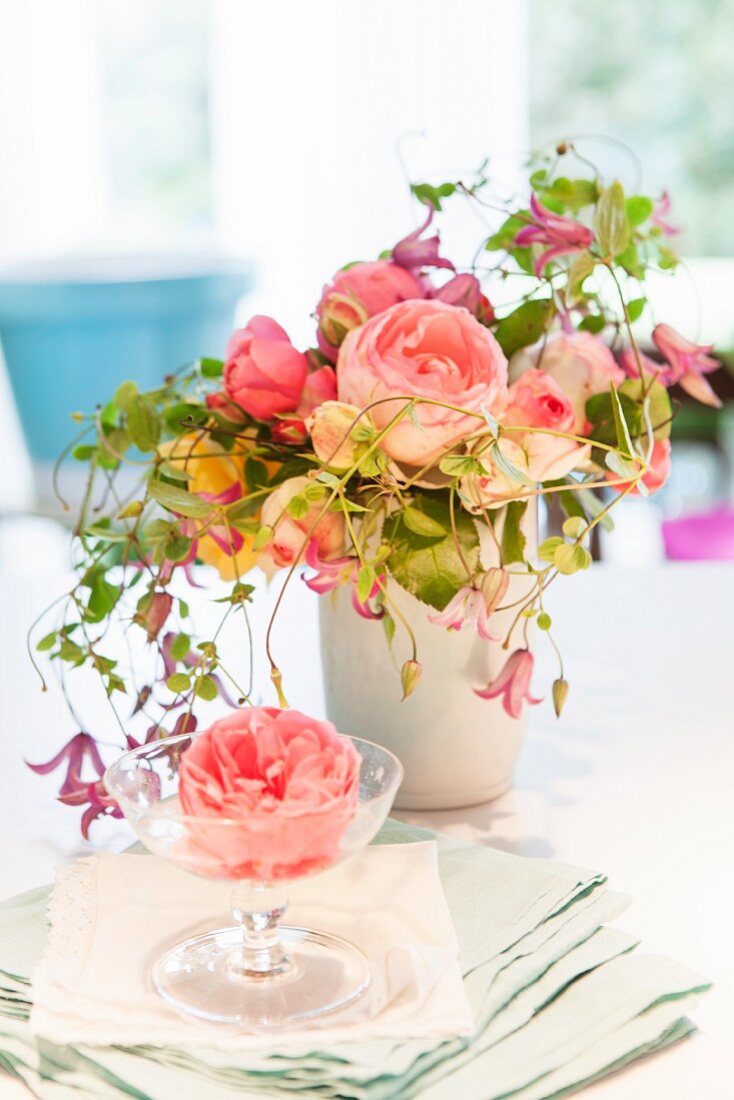 Rosafabene Rosenblüte in Sektschale auf Serviettenstapel und Blumenstrauss mit englischen Duftrosen und Clematis in weisser Vase auf Tisch