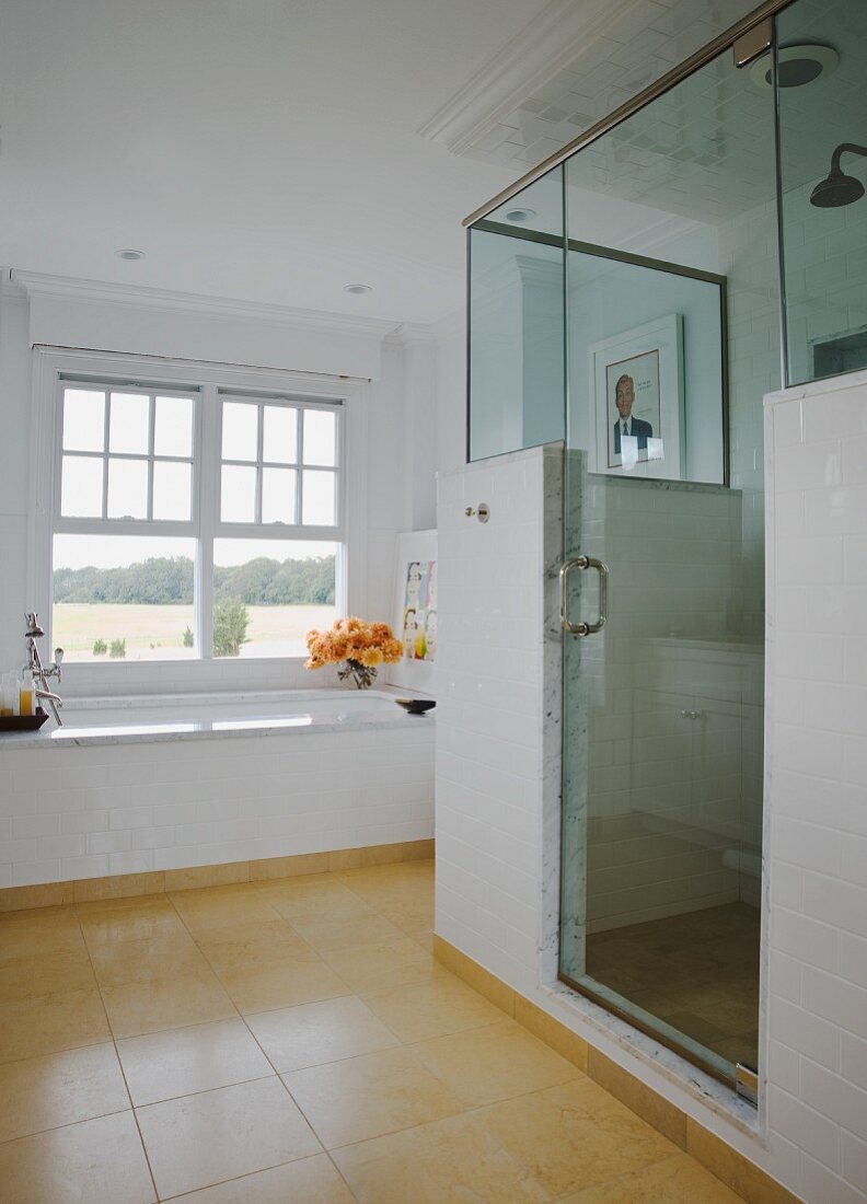 Teilverglaste Duschkabine in Bad mit beigegelben Bodenfliesen; im Hintergrund eine Wanne unter Fenster mit Ausblick
