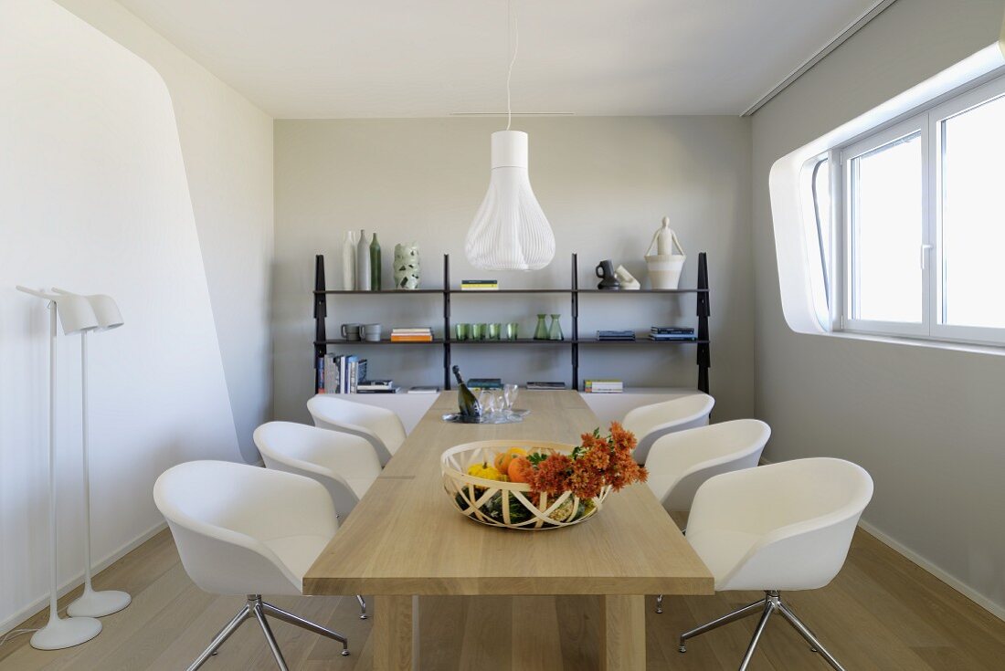 Narrow, pale grey and white dining room with white designer chairs and dining table in solid, pale wood; delicate, black metal shelving against wall