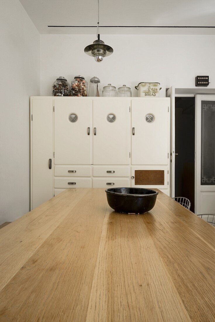 Vintage kitchen cupboard against white wall; pale, solid wood table in foreground