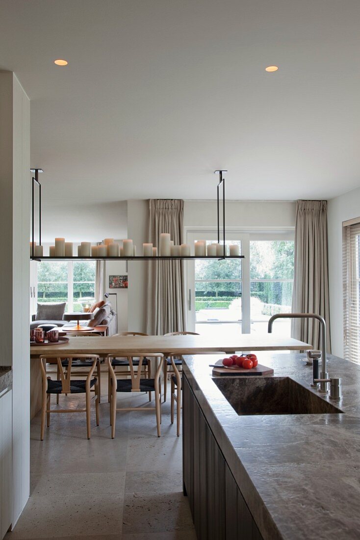 Free-standing kitchen counter with integrated sink in front of dining area with classic wooden chairs, wooden table and collection of white candles on shelf suspended from ceiling