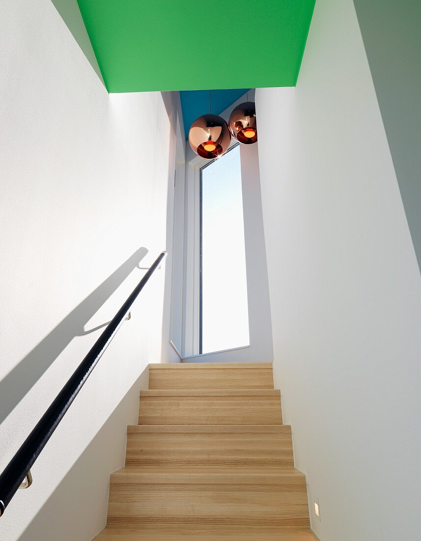 Helle Holztreppe und schwarzer Handlauf in schmalem Treppenhaus, über Podest Pendelleuchten mit kupferfarbenen Schirmen, davor grün gestrichener Deckenbereich