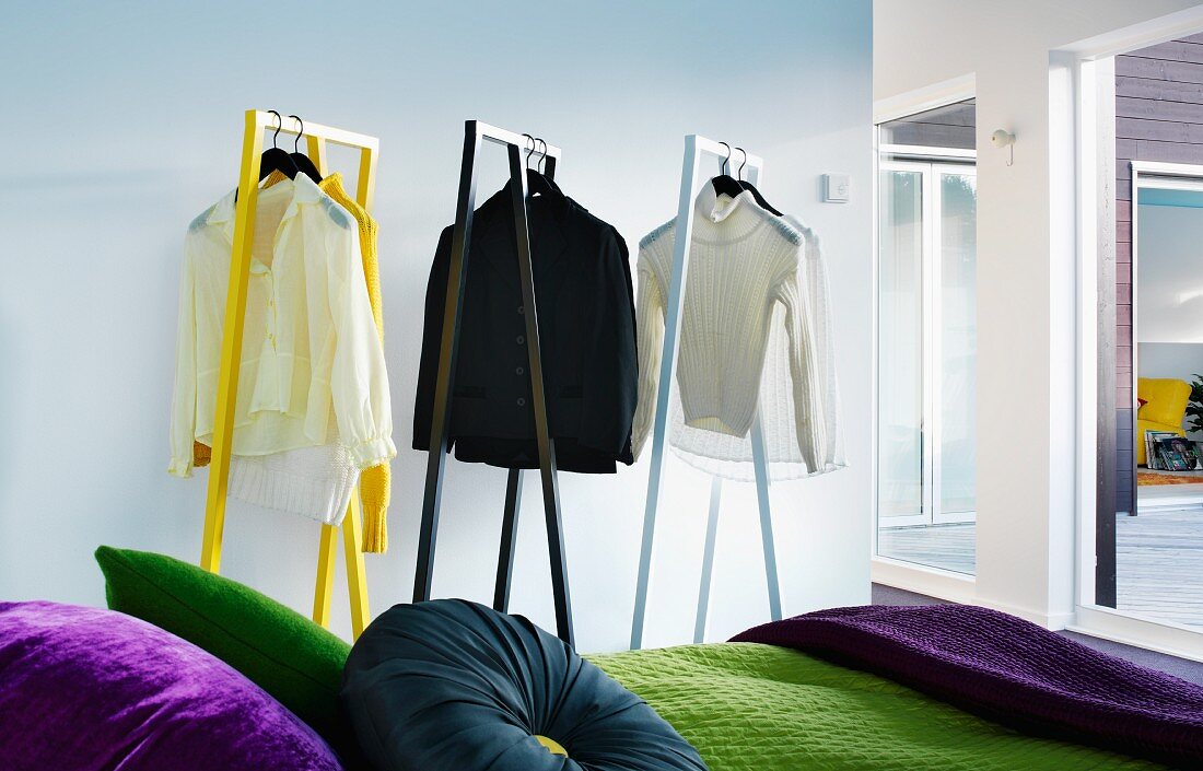 Shirts and jumpers on designer clothes racks behind partially visible bed with scatter cushions of various colours and green bedspread