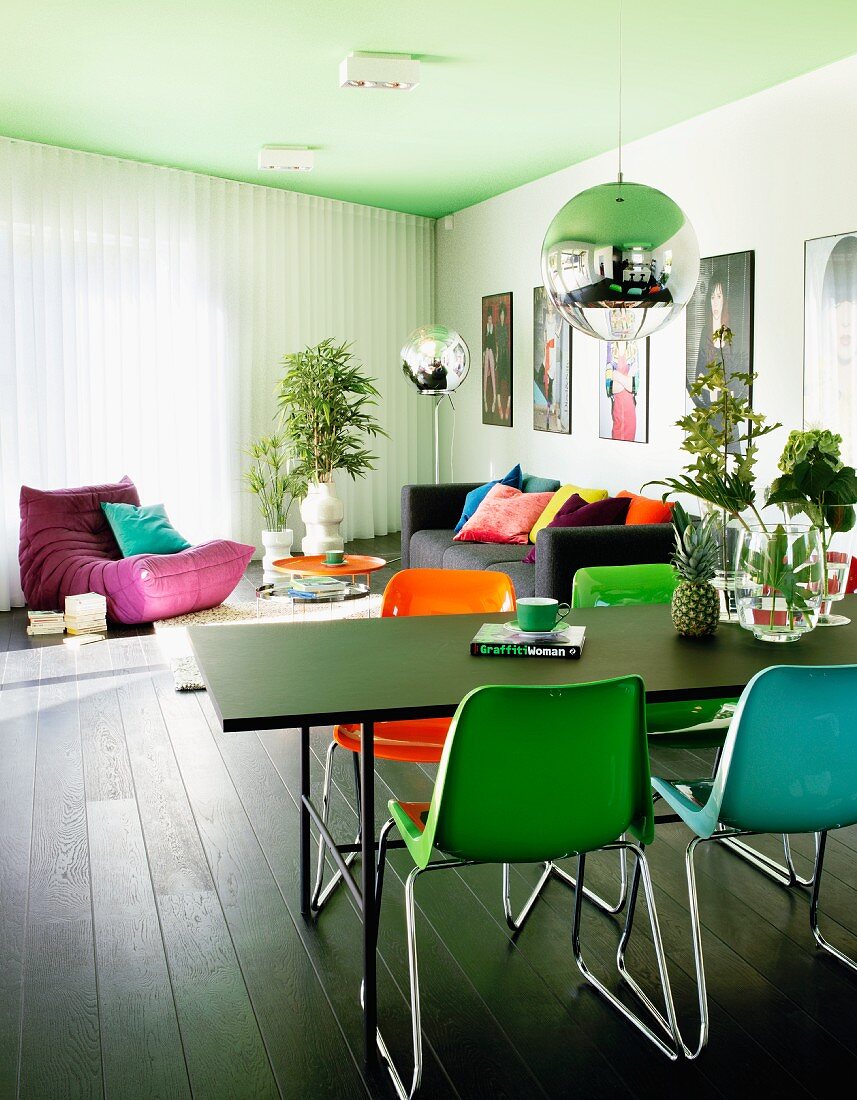 Farbige Kunststoff Stühle an schwarzem Tisch, unter Kugel Pendelleuchte, im Hintergrund Loungebereich, mit Sofa und Sitzsack unter grüngetönter Decke in modernem Wohnraum