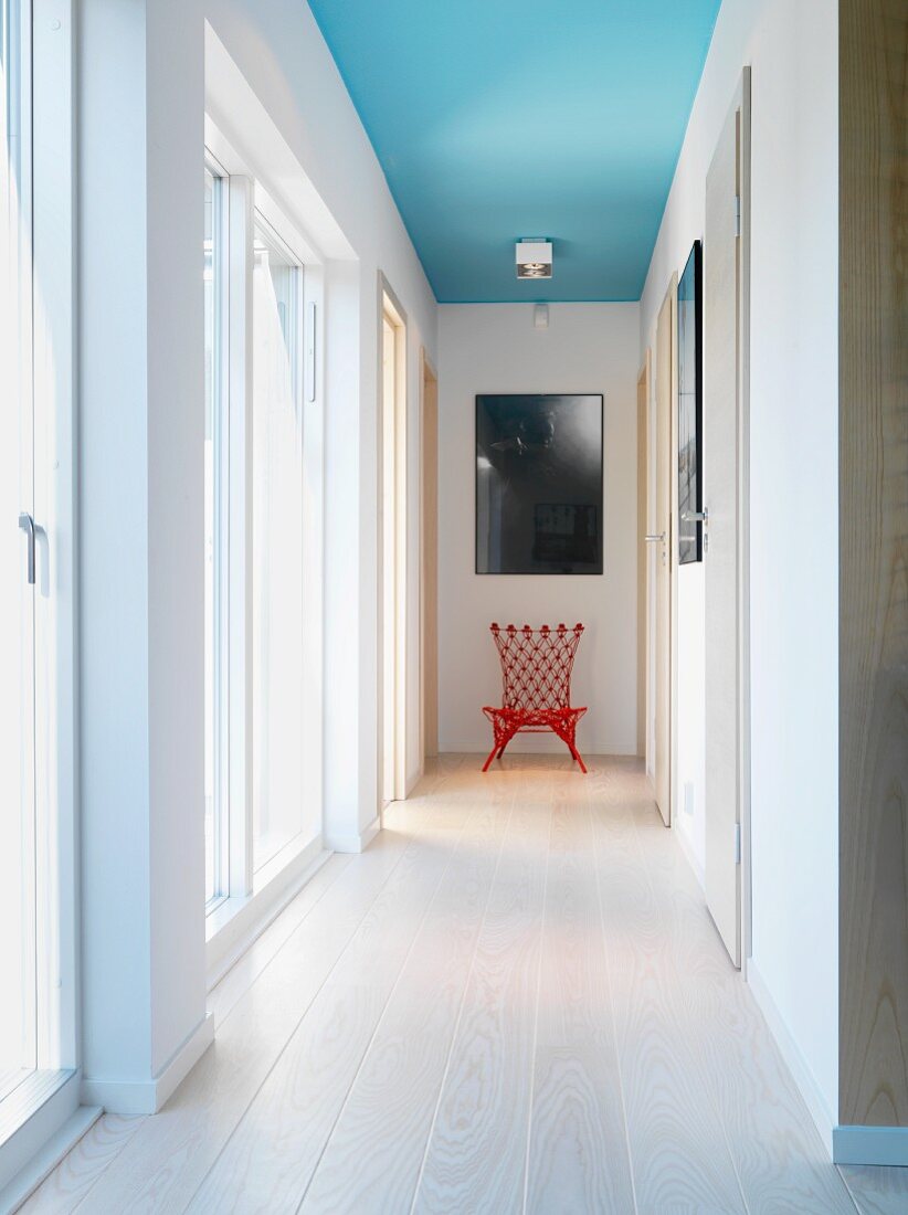Light-flooded hallway with pale wooden floor, red chair below picture on end wall and spotlight on ceiling painted light blue