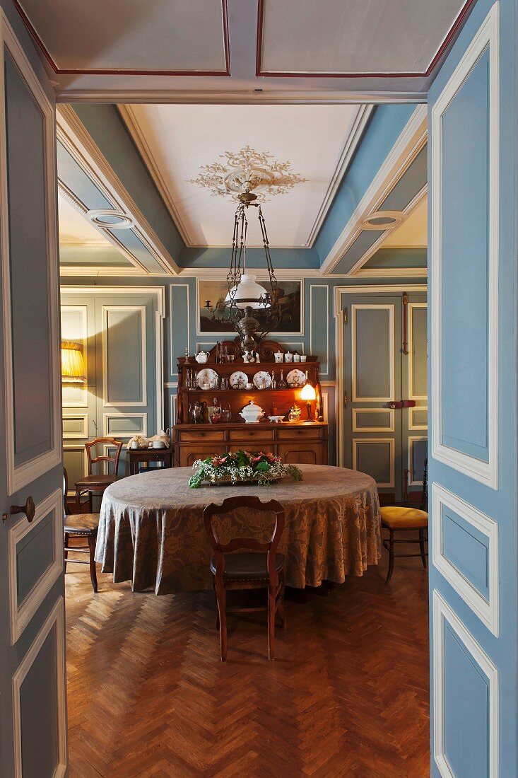 Dining room with blue and white wood panelling, round table, dresser and collection of antique chairs