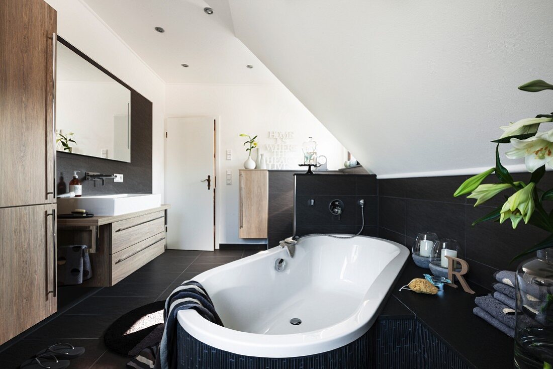 Badewanne diagonal in Raum positioniert unter Dachschräge, an Wand halbhoch braune Fliesen, gegenüber trogartiger Waschtisch und grosser Spiegel an Wand in modernem Bad