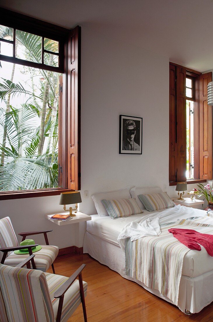 Gemütliches Doppelbett vor Wand, seitlich Sessel vor Fenster mit Holz Innenläden und Blick auf Palmen