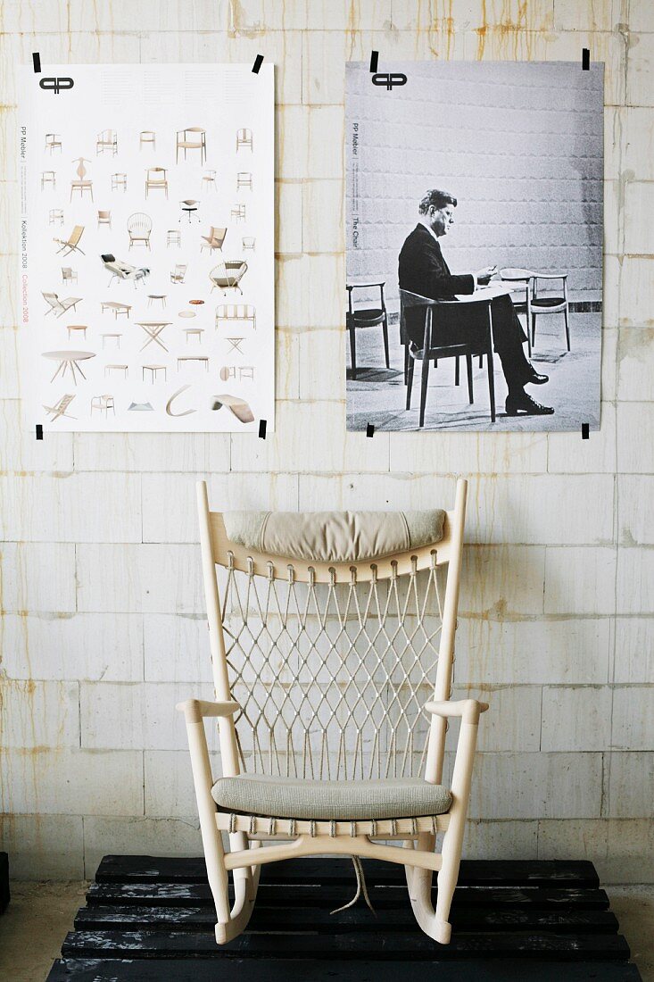 Rocking chair with white-painted wooden frame on wooden pallet below posters taped on unrendered sand-line brick wall