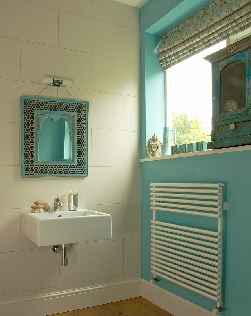 Mirror with ornate, Moroccan frame above modern sink; radiator-towel rack on turquoise wall to one side and antique cabinet on windowsill