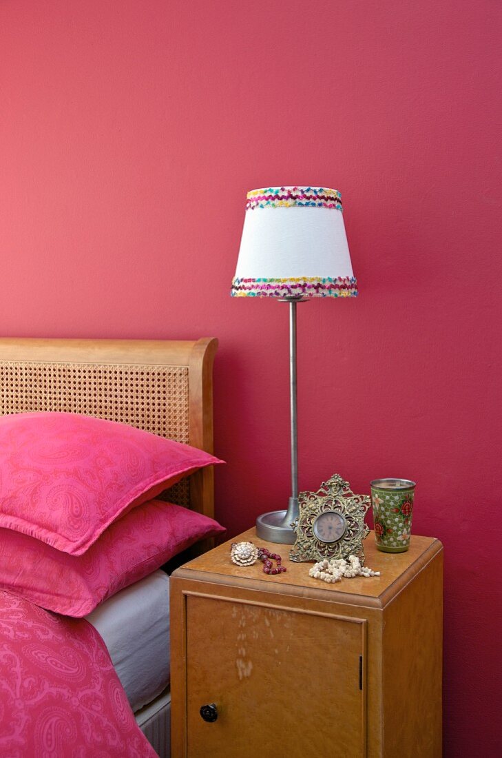 Nachtkästchen mit Lampe vor himbeerroter Wand, daneben Bettwäsche in Pink auf Jugendstil Bett mit Geflecht