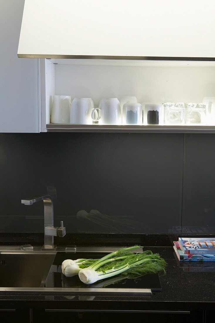 Ausschnitt einer Küchenzeile mit Edelstahl Spülbecken in schwarzer Arbeitsplatte, Spritzschutz aus grau lackiertem Glas, oberhalb offener Hängeschrank mit Innenbeleuchtung