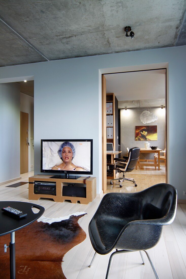 Black retro shell chair, animal skin rug, wooden floor, flatscreen TV on low sideboard and doorway leading to study