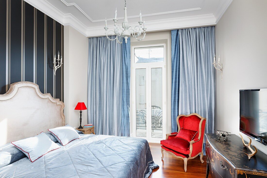 Hellblaue Tagesdecke auf Doppelbett mit geschwungenem Kopfteil, seitlich Nachttischleuchte mit rotem Schirm, im Hintergrund Balkontür mit bodenlangen Vorhängen