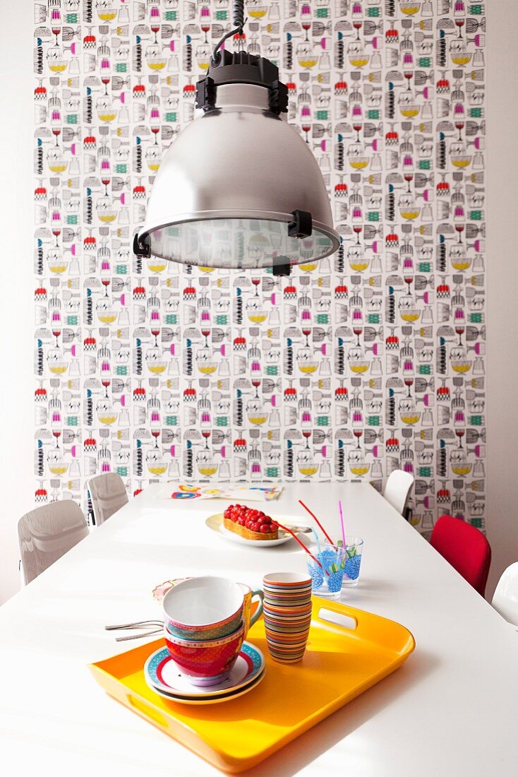 Gelbes Tablett mit Frühstücksgeschirr auf Esstisch, darüber Pendelleuchte, im Hintergrund tapezierte Wand