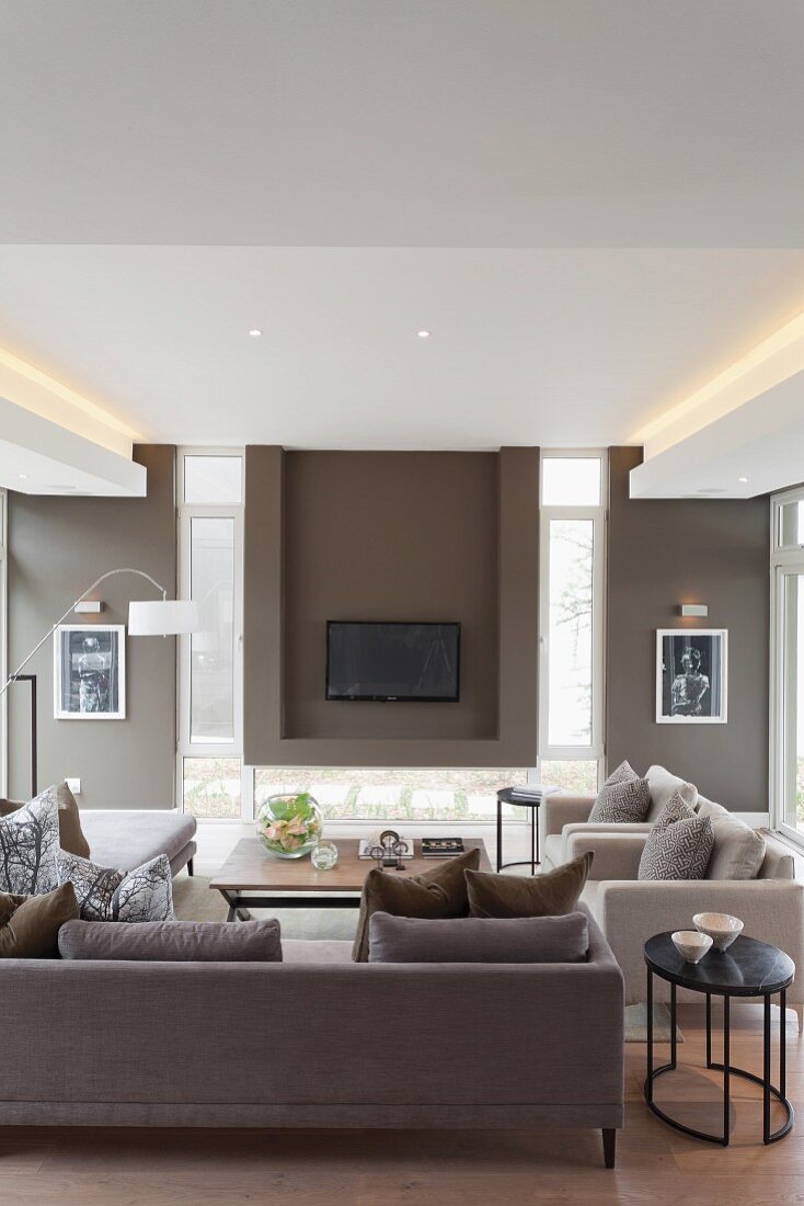 Elegante Sofagarnitur in verschiedenen Brauntönen, gegenüber Wandnische mit aufgehängtem Flachbildschirm zwischen schmalen, hochformatigen Fenstern, indirekte Deckenbeleuchtung