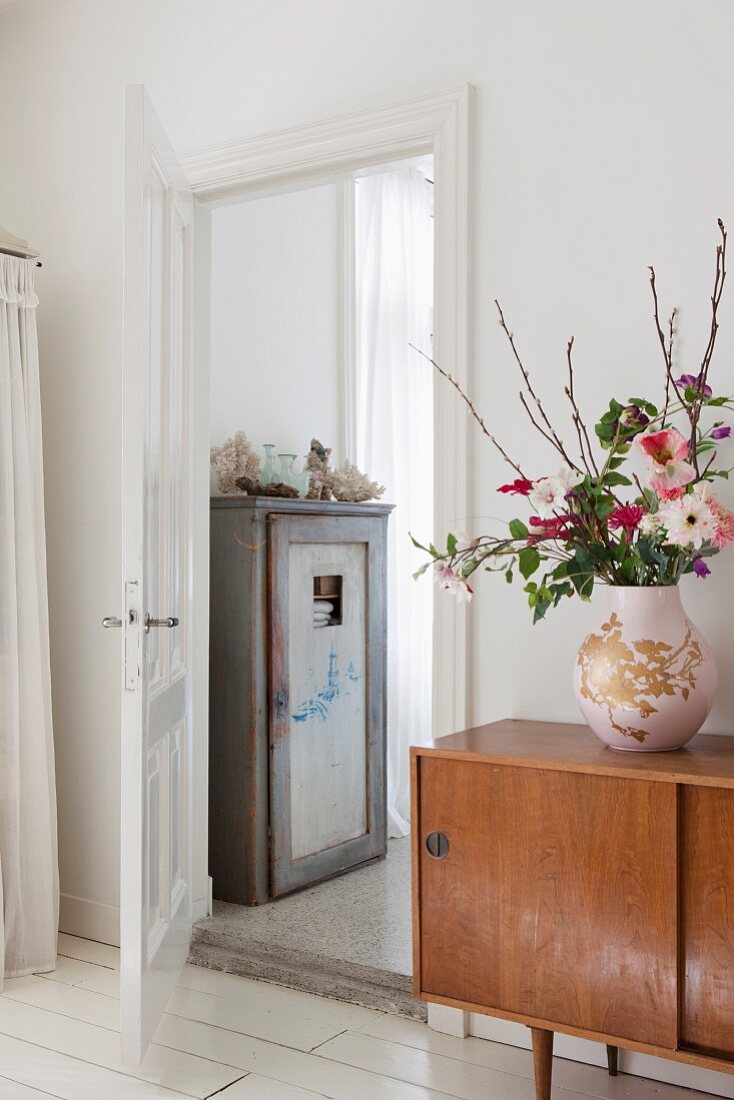 Bulbous vase of flowers on retro sideboard next to open door showing old wooden cupboard in ensuite bathroom