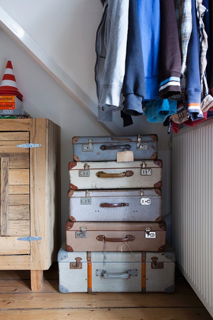 Wäscheaufbewahrung in gestapelten Vintage Koffern im Schlafzimmer, darüber aufgehängte Jacken und Hemden; seitlich ein rustikales Sideboard