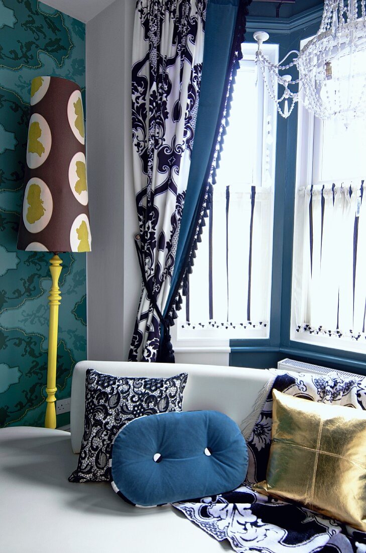 Kissenmix auf Sofa in Fenstererker mit drapiertem Vorhang und Stehleuchte vor Tapetenmuster