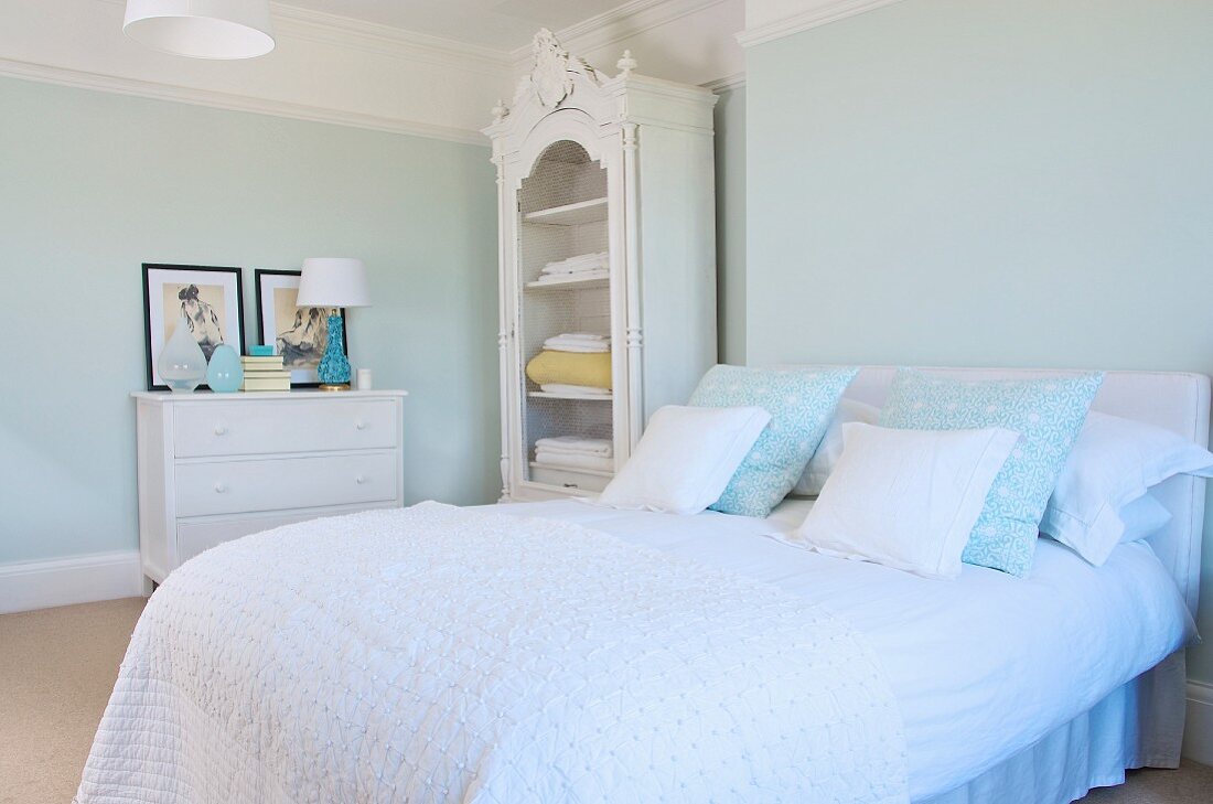 Französisches Bett mit drapierten Kissen und weisser Tagesdecke, weiss lackierter, ländlicher Wäscheschrank mit Schnitzereien in pastellfarbenem Schlafzimmer einer Altbauwohnung