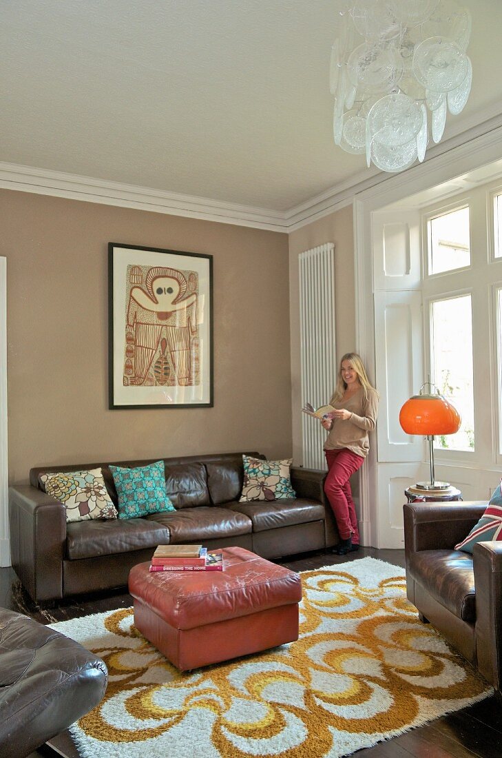 Pink, leather ottoman on patterned rug and woman standing next to brown, leather sofa in traditional interior