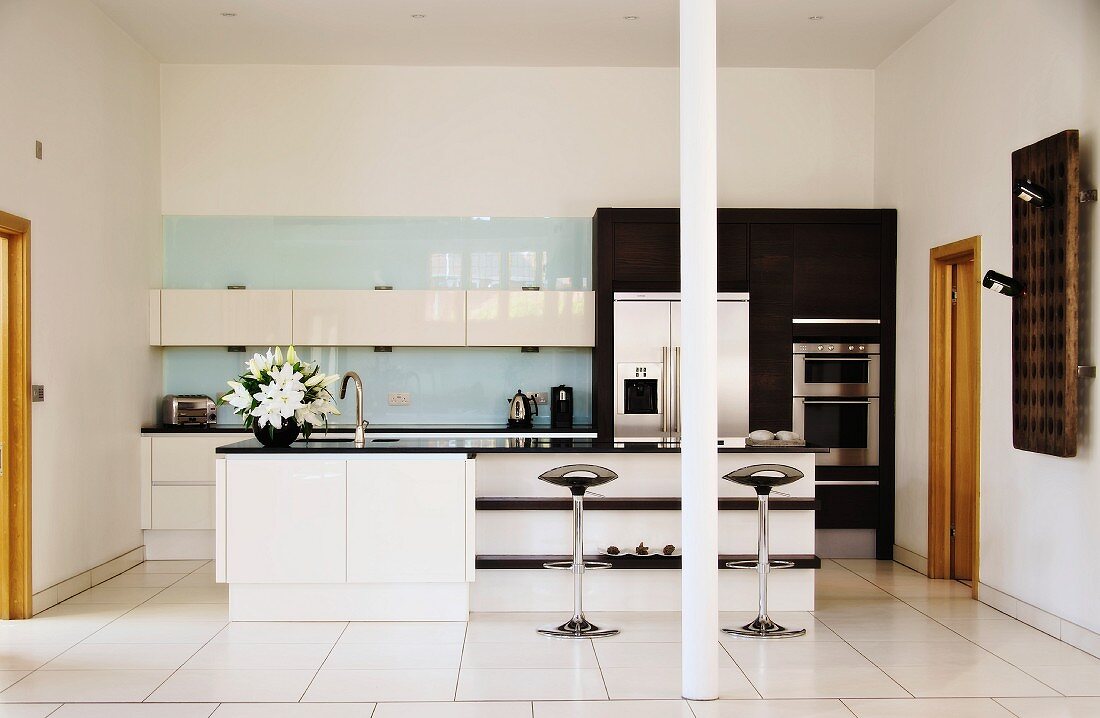 Free-standing counter with shelves and designer bar stools on white tiled floor in open-plan, modern fitted kitchen