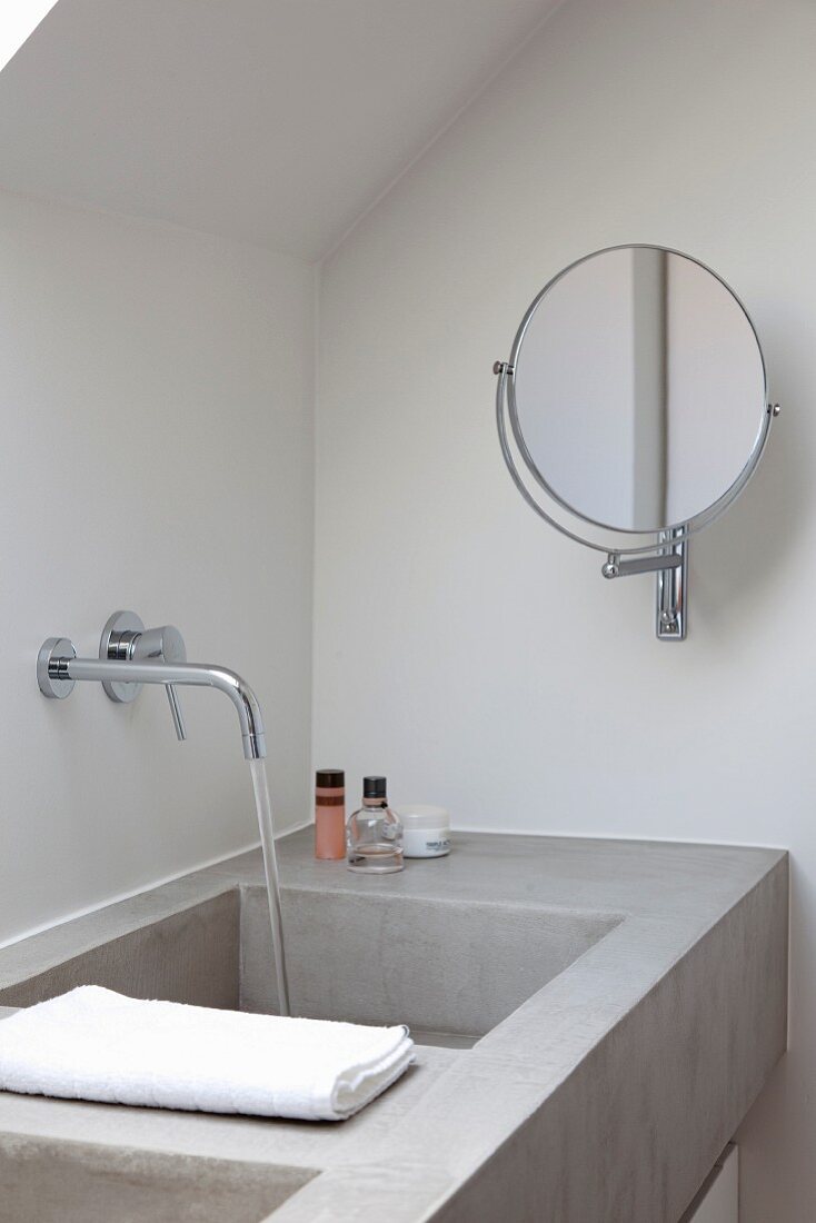 Concrete washstand with water running out of wall-mounted taps and swivelling vanity mirror