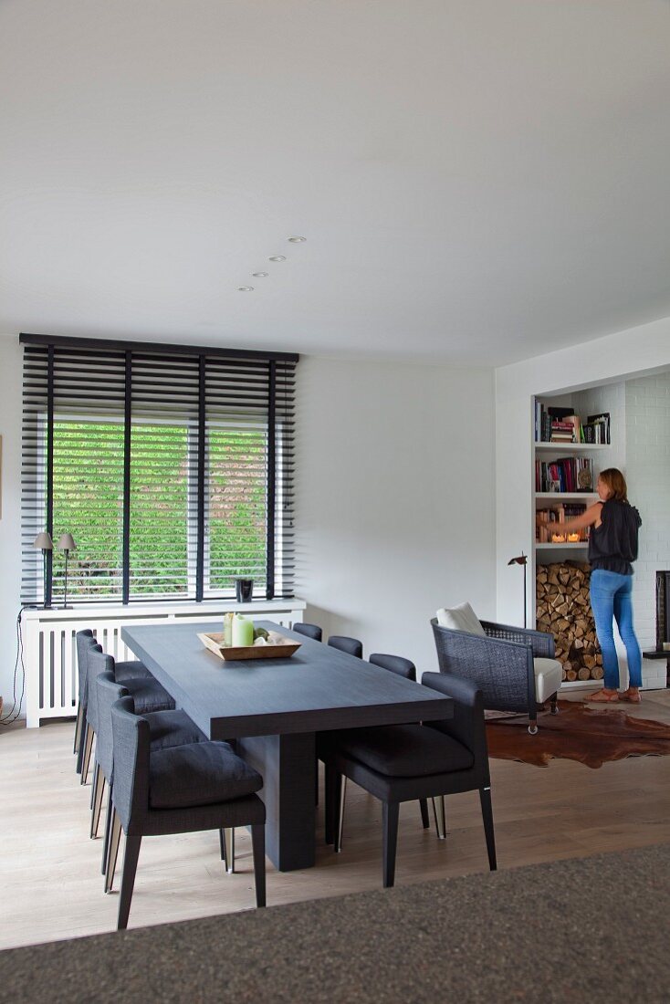 Long table and chairs in slate grey in modern interior; woman in background in front of bookcase with stacked firewood