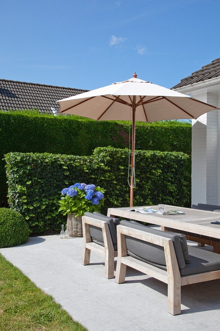 Large parasol and blue hydrangea in planter in front of clipped hedge; dining area with wooden furniture and grey seat cushions